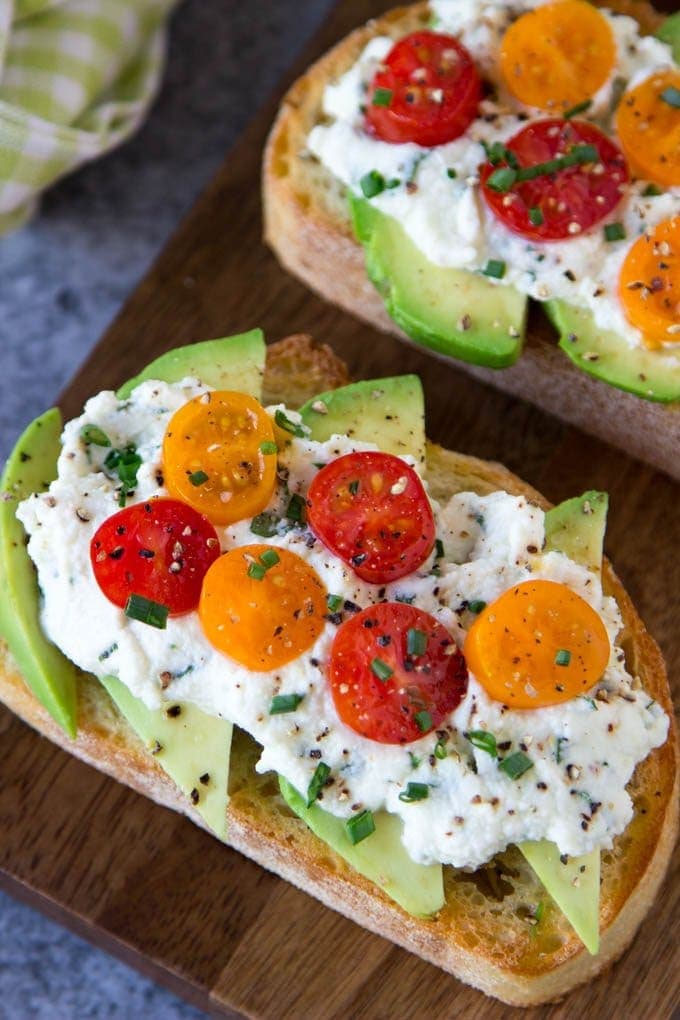 Avocado Toast + Herbed Ricotta & Fresh Tomatoes- Crusty Artisan bread toasted and topped with creamy avocado, herbed ricotta (ricotta with spinach, basil, red pepper flakes and chives) and Fresh Tomatoes. A crowd pleaser and it only takes 5 min. to make.