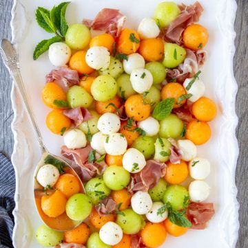 summer melons (honeydew and cantaloupe )balls and mozzarella balls with prosciutto on a white platter with serving spoon