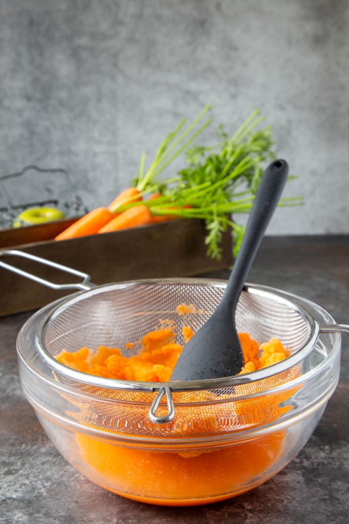 A fine mesh strainer over a large clear bowl. The strainer is filled with the blended carrot apple juice and a rubber spatula is shown in the mesh strainer to push juice through the strainer.